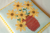 A close-up of a finished cross-stitch piece featuring yellow sunflowers with green stems and a red pot, bordered by yellow and white gingham fabric.