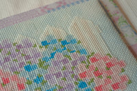 Close-up of a cross-stitch or needlepoint fabric design showing a pastel landscape with lavender, blue, pink, and green stitched flowers on a pale blue and cream background.