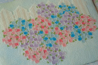 A close-up of a cross-stitch or needlepoint canvas featuring a floral pattern stitched in pastel pink, purple, and blue flowers with small green leaves on a pale blue-green fabric background.
