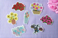 Seven die-cut stickers featuring various colorful floral designs, arranged on a purple and white gingham checkered surface next to a pink rose.
