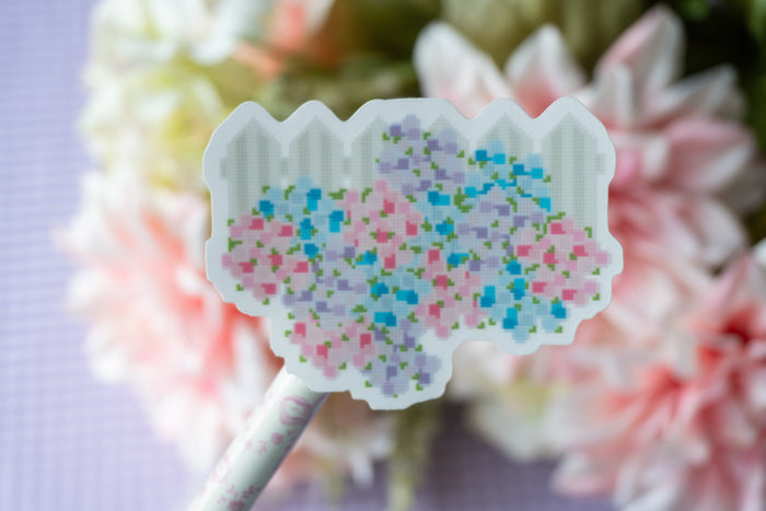 A close-up of a decorative sticker or patch with a pixelated floral pattern in pink, blue, and purple on a white scalloped outline, attached to a patterned stick, set against a soft-focus background of white and pink flowers.
