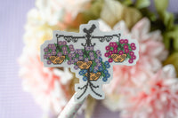 A cross-stitch style decorative sticker or patch featuring two hanging flower baskets with purple and pink blossoms, shown against a blurred background of pale pink and white flowers.