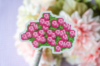 A die-cut sticker featuring a pixel-art style pink flower cluster with green leaves, mounted on a decorative stick and photographed against a soft-focus floral background.