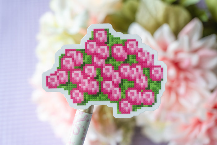 A die-cut sticker featuring a pixel-art style pink flower cluster with green leaves, mounted on a decorative stick and photographed against a soft-focus floral background.