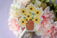 A close-up of a cross-stitch style flower bouquet patch or sticker on a stick, featuring yellow flowers with brown centers in a terracotta pot, set against a blurred background of pink and white flowers.