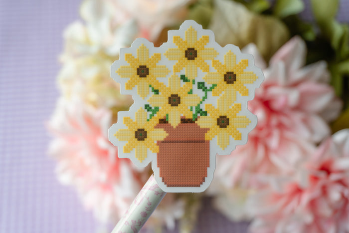 A close-up of a cross-stitch style flower bouquet patch or sticker on a stick, featuring yellow flowers with brown centers in a terracotta pot, set against a blurred background of pink and white flowers.