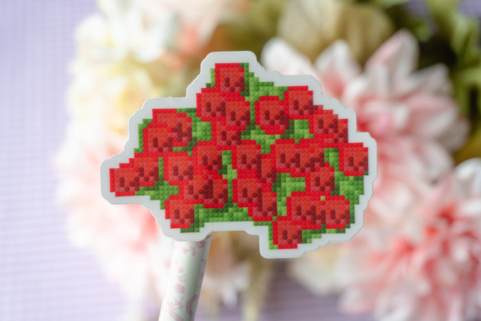 A pixel-art style sticker or patch featuring a bouquet of red flowers with green leaves, mounted on a white stick, set against a soft-focus background of white and pink flowers.