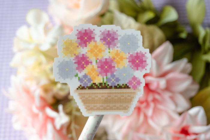 A cross-stitch style flower bouquet patch or sticker on a stick, featuring pink, yellow, and lavender flowers in a beige basket, set against a blurred background of pink and white flowers.