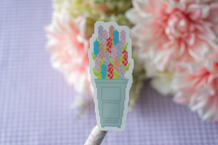 A close-up of a die-cut sticker featuring a pixel-art style bouquet of colorful flowers in a light blue pot, attached to a patterned stick, set against a soft-focus background of pink flowers and a lavender gingham surface.