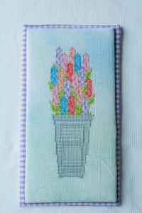 A rectangular needlepoint canvas featuring a colorful bouquet of tulips in a gray pot, bordered by a purple and white gingham pattern, displayed on a white textured fabric background.