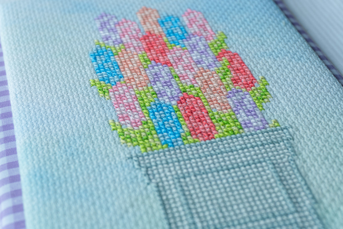 Close-up of a light blue needlepoint canvas featuring a colorful cross-stitch design of flowers in a pot, with visible mesh texture and a purple-and-white striped edge on the left.