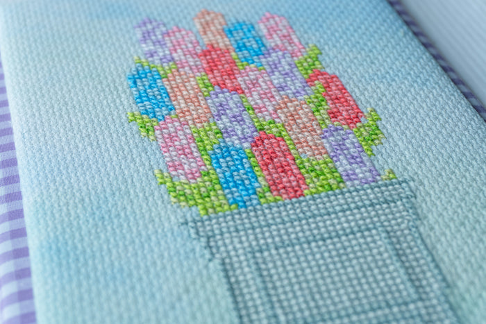 Close-up of a light blue needlepoint canvas featuring a colorful cross-stitch design of flowers in a pot, with visible mesh texture and a purple-and-white striped edge on the left.