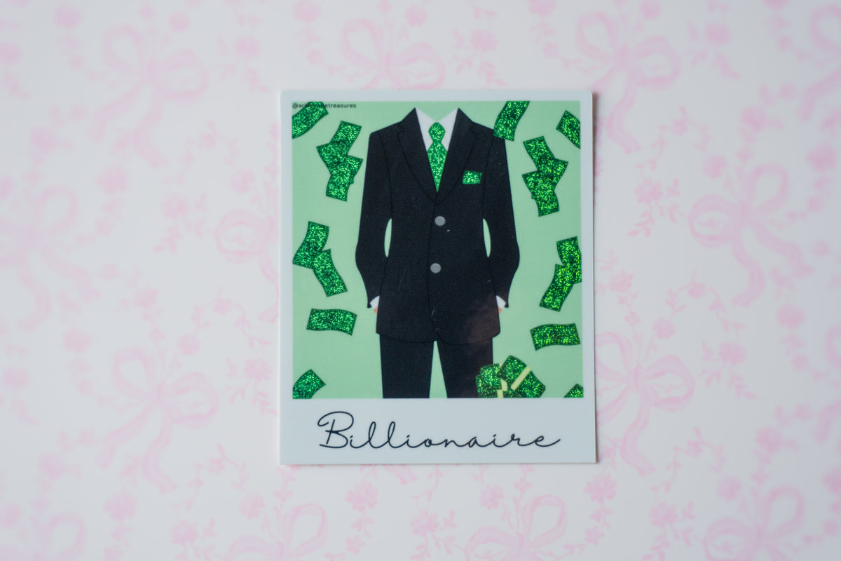 A square glitter polaroid sticker featuring a black suit with green patterned details and scattered green dollar bills on a mint green background, with the word 'Billionaire' written at the bottom. The print is displayed on a light pink patterned surface.