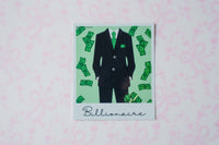 A square glitter polaroid sticker featuring a black suit with green patterned details and scattered green dollar bills on a mint green background, with the word 'Billionaire' written at the bottom. The print is displayed on a light pink patterned surface.