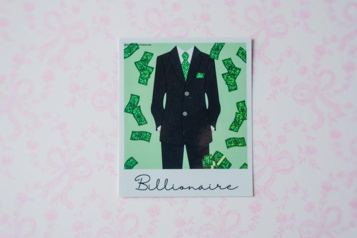 A square glitter polaroid sticker featuring a black suit with green patterned details and scattered green dollar bills on a mint green background, with the word 'Billionaire' written at the bottom. The print is displayed on a light pink patterned surface.