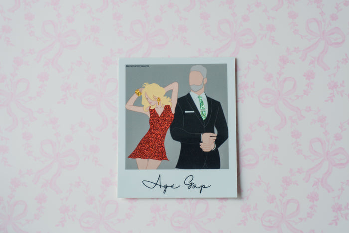 A square glitter polaroid sticker with a white border, featuring a stylized illustration of a blonde woman in a red dress next to a man in a black suit with a green patterned tie. The text 'Age Gap' is written in black script at the bottom.
