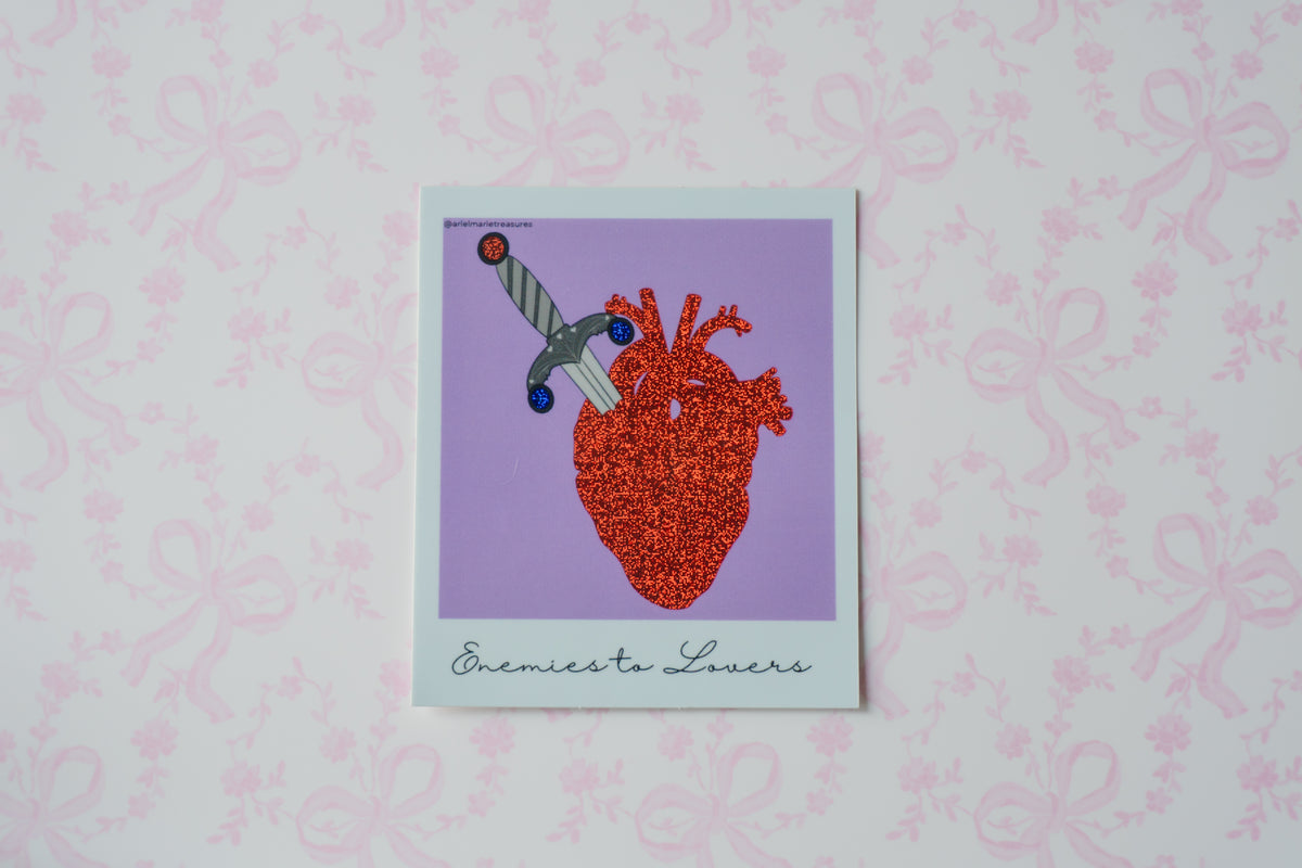 A square Polaroid glitter sticker with a white border featuring a stylized anatomical heart in red and orange tones with a dagger, set against a lavender background. The print is titled 'Enemies Lovers' in cursive at the bottom, displayed on a light pink patterned background.