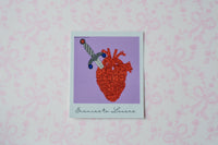 A square Polaroid glitter sticker with a white border featuring a stylized anatomical heart in red and orange tones with a dagger, set against a lavender background. The print is titled 'Enemies Lovers' in cursive at the bottom, displayed on a light pink patterned background.