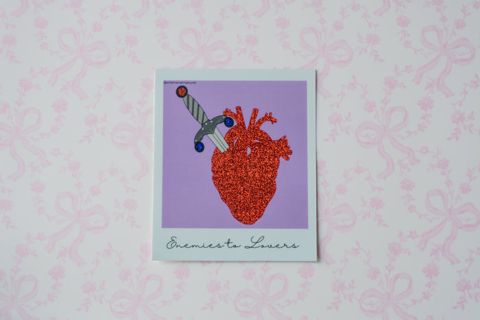 A square Polaroid glitter sticker with a white border featuring a stylized anatomical heart in red and orange tones with a dagger, set against a lavender background. The print is titled 'Enemies Lovers' in cursive at the bottom, displayed on a light pink patterned background.
