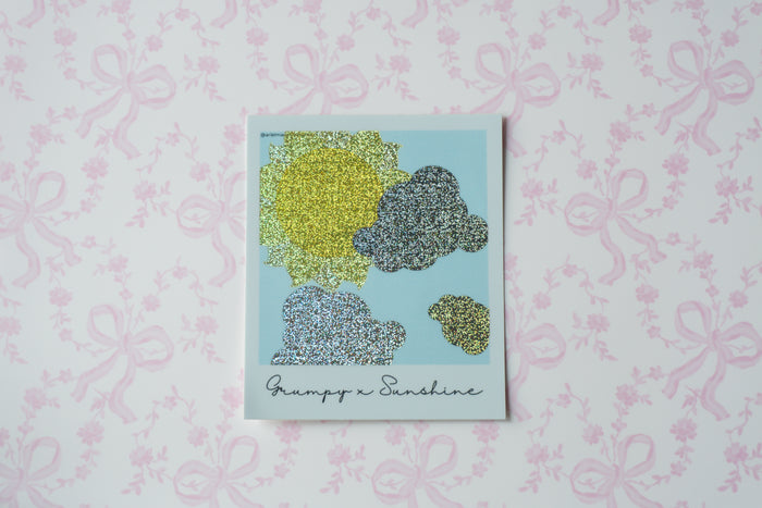 A square glitter polaroid sticker  titled 'Grumpy x Sunshine' featuring abstract cloud-like shapes in yellow, gray, and blue-gray on a light blue background, displayed on a pink patterned wallpaper.
