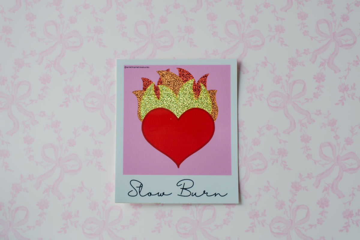 A square glitter sticker featuring a red heart with glittery gold flames above it, set against a pink background with the words 'Slow Burn' written at the bottom. The print is placed on a white surface with a light pink bow-patterned background.