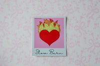 A square glitter sticker featuring a red heart with glittery gold flames above it, set against a pink background with the words 'Slow Burn' written at the bottom. The print is placed on a white surface with a light pink bow-patterned background.