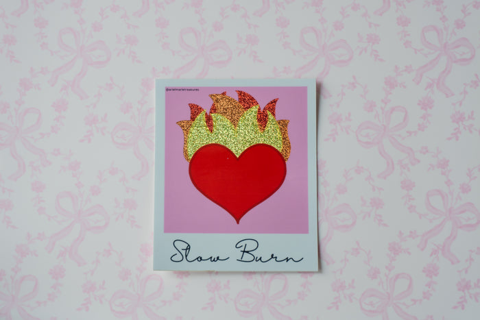 A square glitter sticker featuring a red heart with glittery gold flames above it, set against a pink background with the words 'Slow Burn' written at the bottom. The print is placed on a white surface with a light pink bow-patterned background.