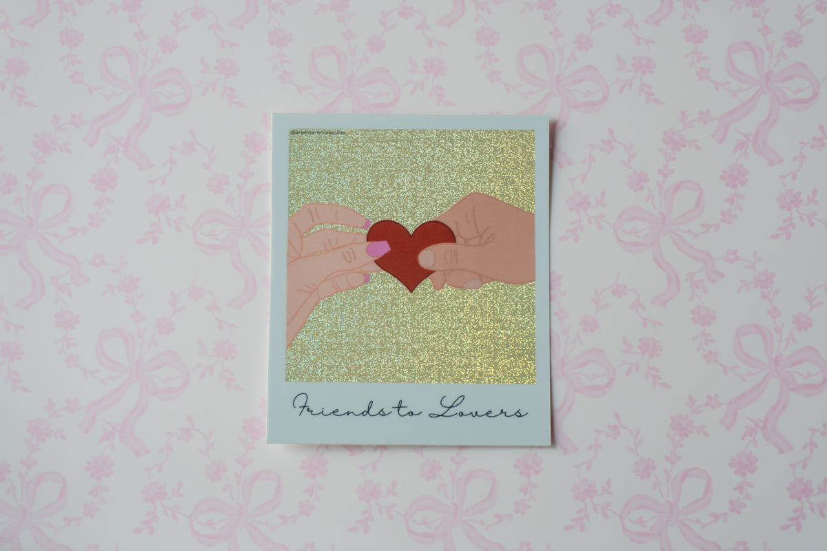 A square polaroid sticker with a white border, featuring an illustration of two hands holding a red heart against a glittery gold background, with the text 'Friends to Lovers' printed at the bottom. The card is displayed on a light pink patterned surface.