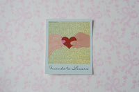A square polaroid sticker with a white border, featuring an illustration of two hands holding a red heart against a glittery gold background, with the text 'Friends to Lovers' printed at the bottom. The card is displayed on a light pink patterned surface.