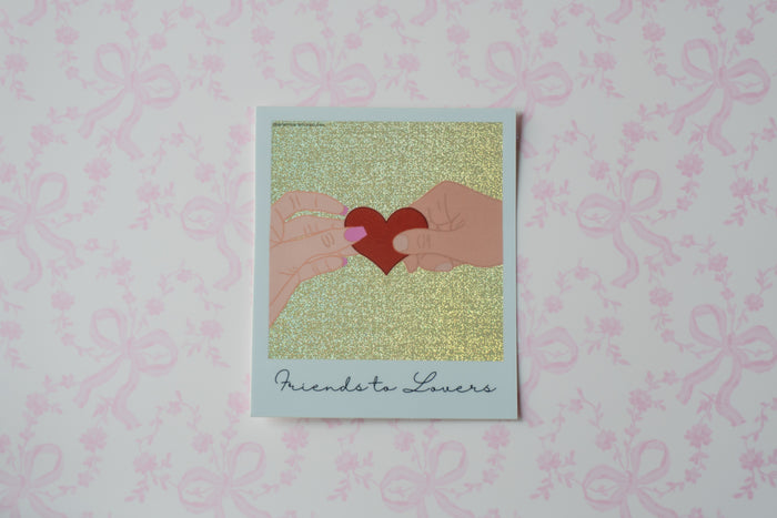 A square polaroid sticker with a white border, featuring an illustration of two hands holding a red heart against a glittery gold background, with the text 'Friends to Lovers' printed at the bottom. The card is displayed on a light pink patterned surface.
