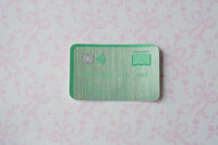 A green metallic credit card sticker with a chip and wireless icon, placed on a light pink patterned background.