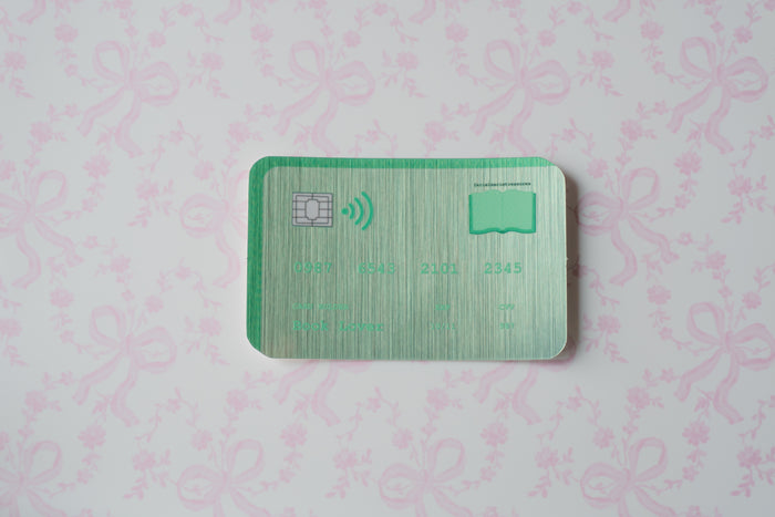 A green metallic credit card sticker with a chip and wireless icon, placed on a light pink patterned background.