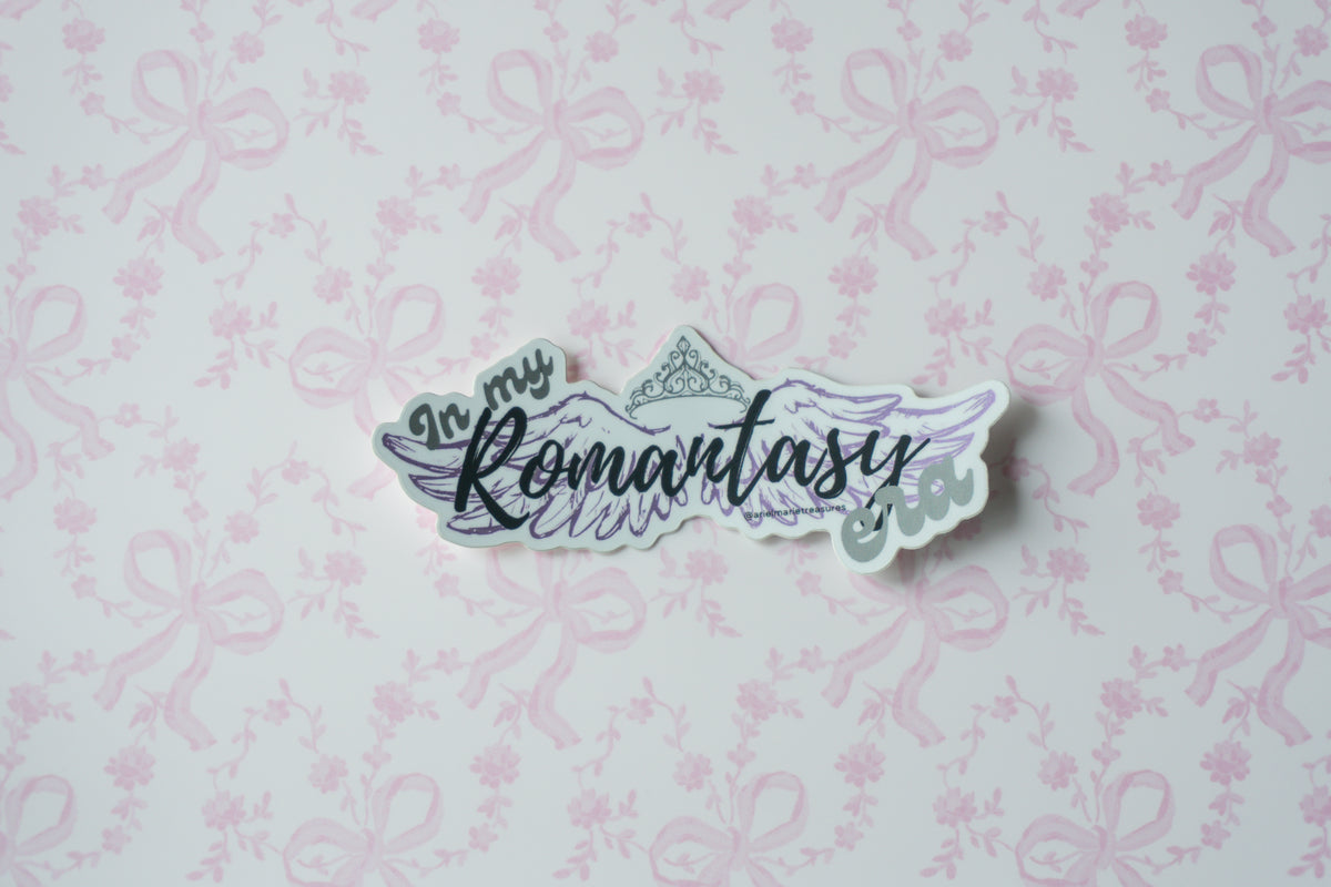 A die-cut sticker with the phrase 'In my Romantasy Fandom' in stylized lettering, featuring wings and a crown illustration, placed on a light pink patterned background.