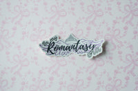 A die-cut sticker with the phrase 'In my Romantasy Fandom' in stylized lettering, featuring wings and a crown illustration, placed on a light pink patterned background.