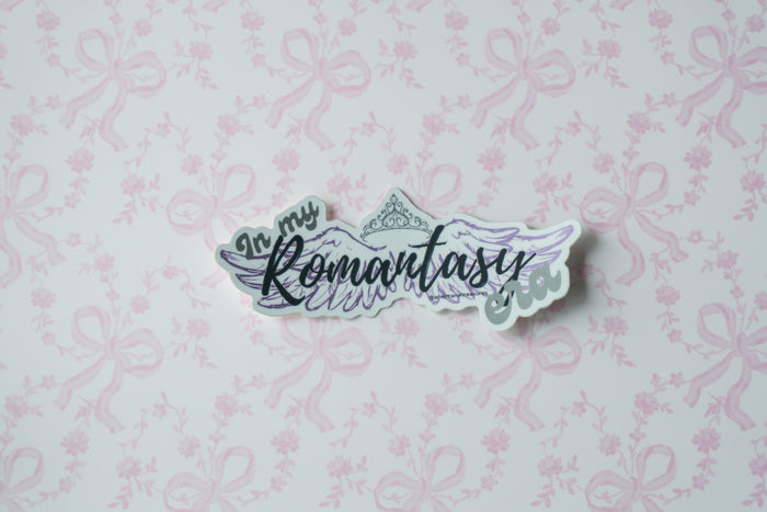 A die-cut sticker with the phrase 'In my Romantasy Fandom' in stylized lettering, featuring wings and a crown illustration, placed on a light pink patterned background.