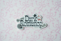 A die-cut sticker with the text 'In my Dark Romance era' in stylized lettering, featuring a small witch hat and a ghost illustration, displayed on a light pink patterned background.