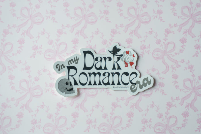 A die-cut sticker with the text 'In my Dark Romance era' in stylized lettering, featuring a small witch hat and a ghost illustration, displayed on a light pink patterned background.