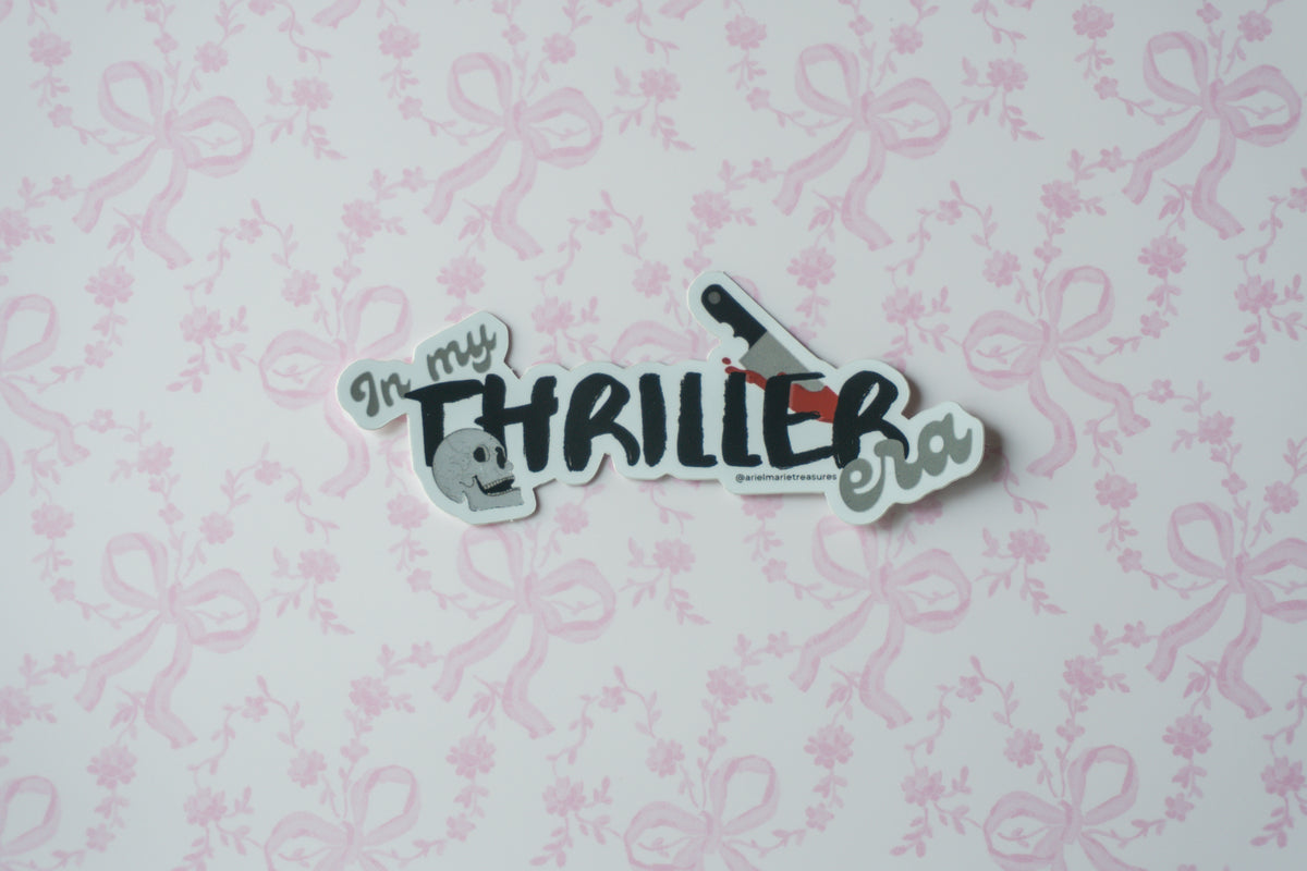A die-cut sticker with the text 'In my THRILLER era!' and small illustrations, placed on a light pink floral wallpaper background.