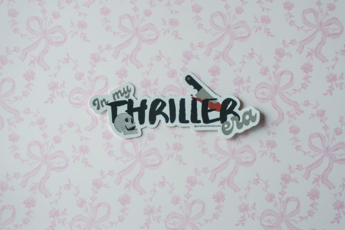 A die-cut sticker with the text 'In my THRILLER era!' and small illustrations, placed on a light pink floral wallpaper background.
