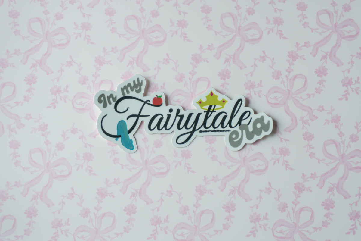 A die-cut sticker with the phrase 'In my Fairytale way' in stylized script, featuring a small red strawberry, blue shoe, and gold crown, displayed on a white background with a light pink floral bow pattern.