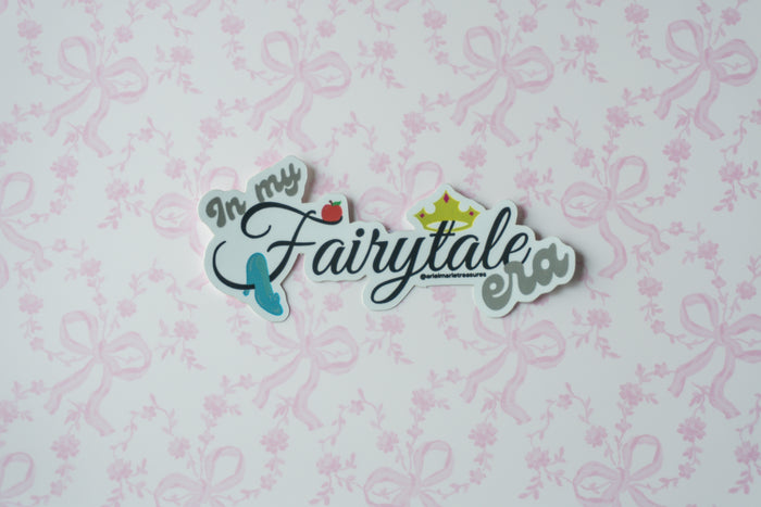 A die-cut sticker with the phrase 'In my Fairytale way' in stylized script, featuring a small red strawberry, blue shoe, and gold crown, displayed on a white background with a light pink floral bow pattern.