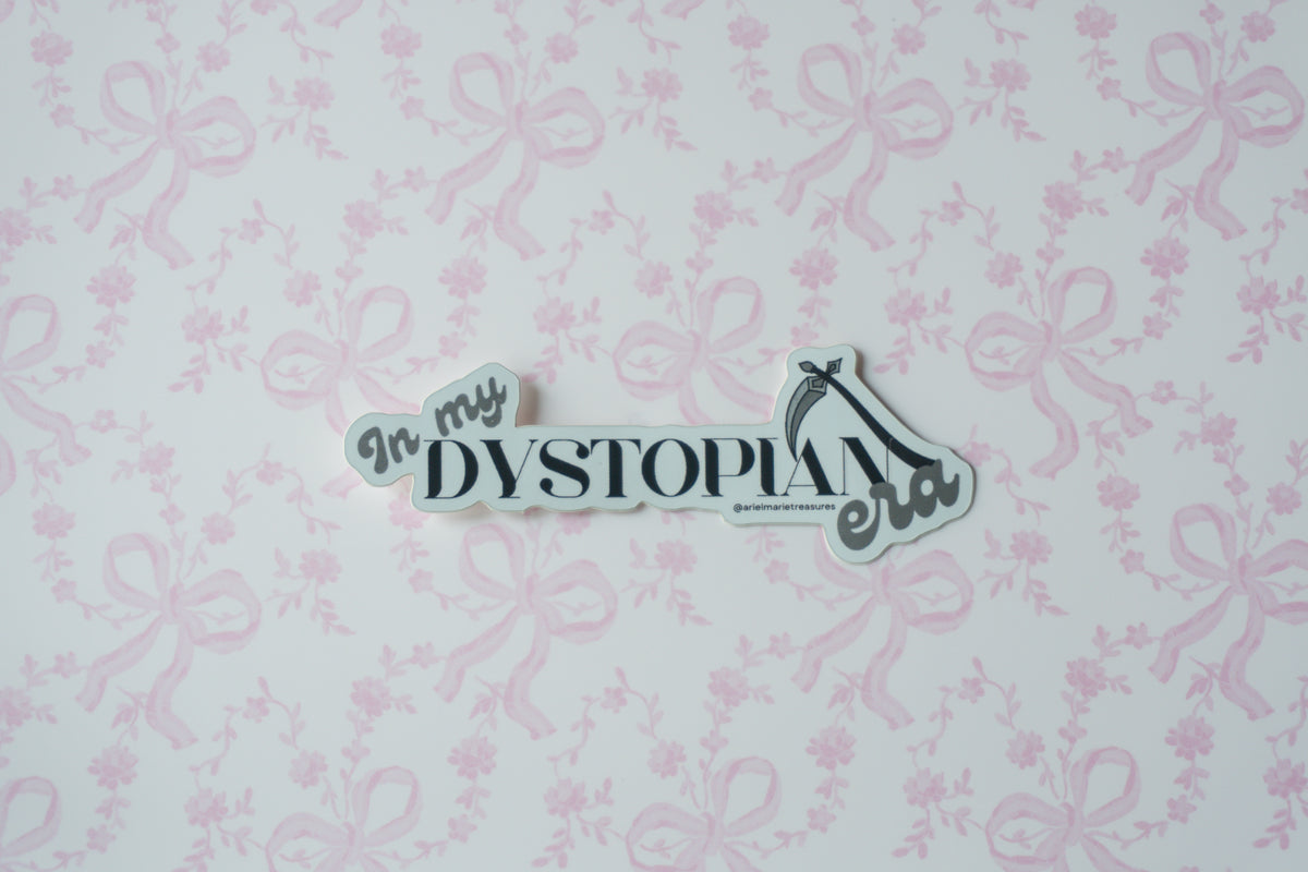 A die-cut sticker with the text 'In my DYSTOPIA era' in black script and serif fonts, featuring a high-heel shoe icon, displayed on a light pink and white patterned background.