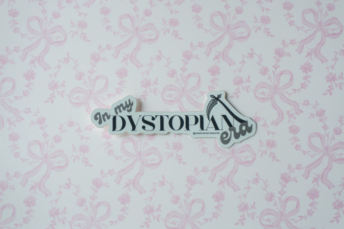 A die-cut sticker with the text 'In my DYSTOPIA era' in black script and serif fonts, featuring a high-heel shoe icon, displayed on a light pink and white patterned background.