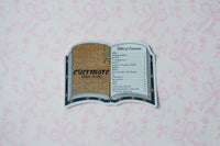 A die-cut sticker shaped like an open book displaying 'evermore (album-cover)' with a gold glittery cover and a black-and-white table of contents page, placed on a light pink patterned background.