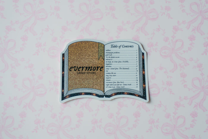 A die-cut sticker shaped like an open book displaying 'evermore (album-cover)' with a gold glittery cover and a black-and-white table of contents page, placed on a light pink patterned background.