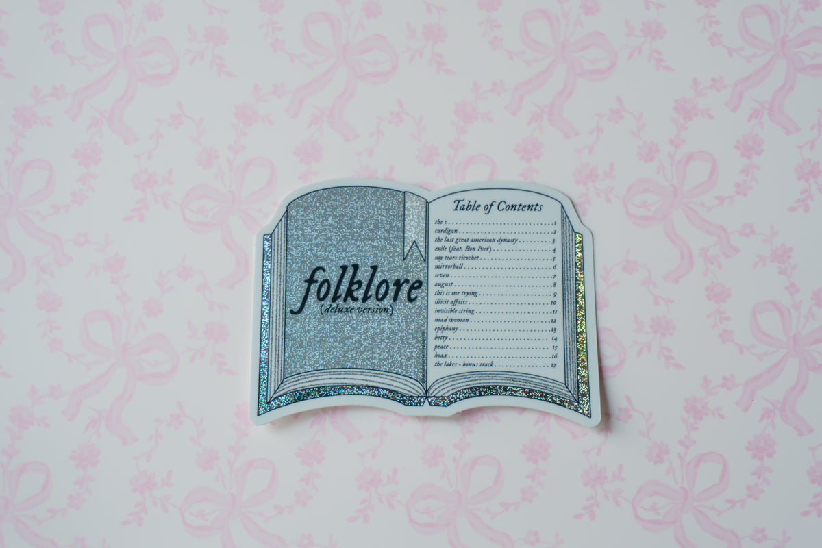 A die-cut sticker shaped like an open book titled 'folklore ' with a table of contents page, placed on a light pink patterned background.