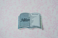 A die-cut sticker shaped like an open book titled 'folklore ' with a table of contents page, placed on a light pink patterned background.