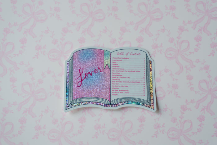 A die-cut sticker shaped like an open book, featuring a glittery pink and blue cover with the word 'loves' in script, and a printed table of contents on the right page. The sticker is displayed on a light pink background with a matching bow pattern.