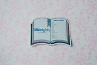 A die-cut sticker shaped like an open book, featuring the title 'Midnights' and a table of contents, displayed on a light pink patterned background.