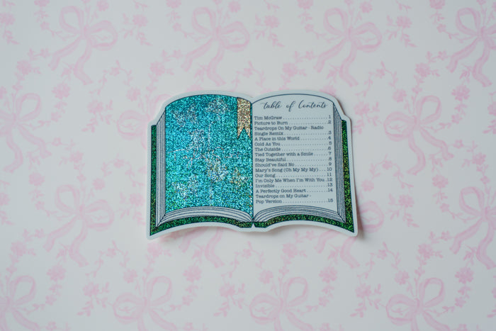 A decorative open book-shaped sticker with glittery turquoise pages and a printed table of contents design, centered on a pale pink patterned background.